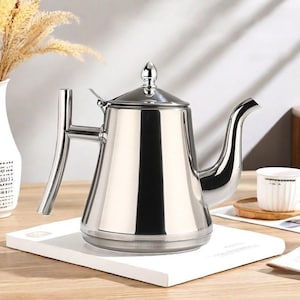 Puede incluir: Una tetera de acero inoxidable pulido con un pico curvo y un asa. La tetera tiene una tapa con un adorno decorativo. Se encuentra sobre una superficie blanca, lo que sugiere un entorno de cocina o comedor. La tetera está diseñada para preparar té.