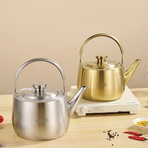 May include: Two stainless steel and gold-colored kettles with arched handles and spouts. The kettles are on a wooden surface with a white heating element. Spices and chili peppers are also visible.