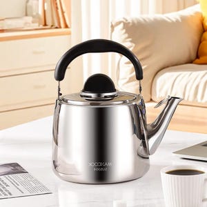 May include: A stainless steel kettle with a black handle and lid. The kettle has a shiny, reflective surface and a spout for pouring. The brand name "MAXCOOK" is printed on the side. A cup of coffee and a laptop are in the background.