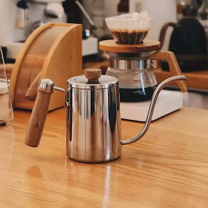 Op de afbeelding: Een roestvrijstalen zwanenhalsketel met een houten handvat en knop staat op een houten oppervlak. De lange, gebogen tuit zorgt voor nauwkeurig schenken. Andere koffiezetapparatuur is zichtbaar op de achtergrond.
