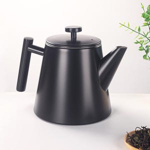 May include: A matte black teapot with a cylindrical body, a curved spout, and a handle. The lid has a small, round knob. The teapot is set against a white background with a hint of greenery and a wooden spoon with tea leaves.