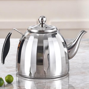 Puede incluir: Una tetera brillante de acero inoxidable con un cuerpo redondeado y un pico curvo. La tetera tiene un asa negra y un pequeño pomo redondo en la tapa. La superficie refleja la luz. Diseñada para uso en estufa.