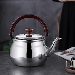 May include: A stainless steel tea kettle with a shiny, reflective surface. It features a dark brown, arched handle and a matching knob on the lid. The spout is curved, and the kettle sits on a dark surface.