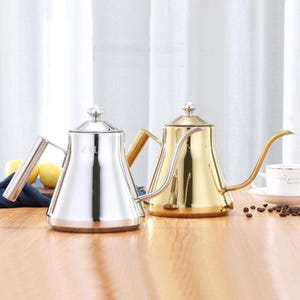 Può includere: Due bollitori a collo di cigno in acciaio inossidabile, uno argentato e uno dorato, su una superficie di legno. I bollitori hanno beccucci lunghi e curvi e piccoli coperchi decorativi. Una tazza di caffè e chicchi di caffè sullo sfondo.