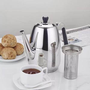 May include: A shiny, stainless steel tea kettle with a black handle and lid, next to a white teacup and saucer. A tea infuser and a plate of bread rolls are also present. The kettle has a built-in thermometer.