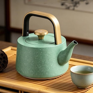 May include: A speckled green ceramic teapot with a gold-coloured handle and lid knob. The spout is curved, and a matching teacup sits nearby on a wooden tray. The teapot has a modern design.
