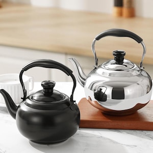 Puede incluir: Dos teteras de acero inoxidable con asas y tapas negras. Una es negra, la otra es plateada brillante. Ambas tienen forma redondeada y un pico para verter. Están sobre una tabla de cortar de madera.