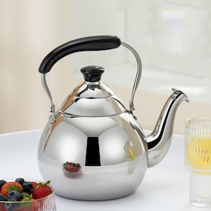 May include: A polished stainless steel kettle with a black handle and lid. The kettle features a curved spout and a rounded body. A bowl of berries and a glass of liquid are also visible in the image.
