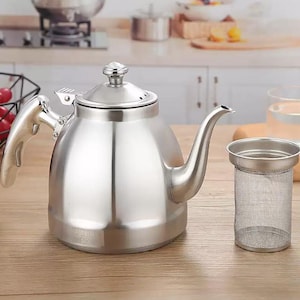 May include: A polished stainless steel teapot with a curved spout and a separate tea infuser. The teapot has a shiny, reflective surface and a handle. The infuser is a cylindrical mesh basket. The teapot and infuser are on a wooden surface.
