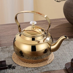 May include: A shiny gold-colored stainless steel teapot with a curved handle and spout. The lid is topped with a clear, faceted knob. The teapot sits on a round, woven coaster, suggesting a tea-drinking theme.
