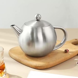 Puede incluir: Una tetera de acero inoxidable con cuerpo redondo, asa curvada y boquilla. La tapa tiene un pequeño pomo redondo. La tetera está sobre una tabla de cortar de madera. La tetera está diseñada para preparar té.