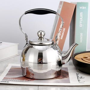 May include: A shiny, stainless steel tea kettle with a black handle and a small, round knob on the lid. The kettle has a curved spout and a large handle. The kettle is sitting on a white surface.