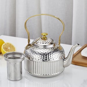 May include: A silver tea kettle with a gold handle and lid finial. The kettle has a textured body and a separate stainless steel tea infuser. A lemon half is visible in the background. The kettle is designed for brewing tea.