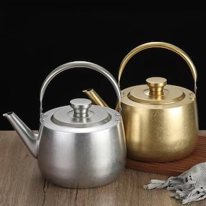 May include: Two teapots, one silver and one gold, are displayed. The silver teapot has a curved handle and a spout. The gold teapot has a similar design. Both teapots have a lid with a small knob. The background is dark.
