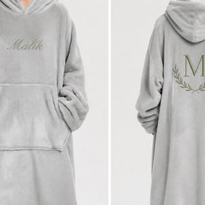 Pode incluir: Um cobertor com capuz cinza claro com um grande bolso frontal. A frente apresenta o nome "Malik" bordado em uma fonte cursiva. A parte de trás tem um "M" bordado dentro de uma coroa de louros.
