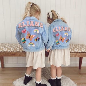 Op de afbeelding: Twee jonge meisjes dragen lichtblauwe spijkerjacks met de naam "ELIANE" in roze letters op de rug. De jacks zijn versierd met kleurrijke patches, waaronder vlinders, regenbogen en teddyberen. Ze dragen beige jurken, zwarte laarzen en witte sokken.
