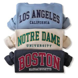 Puede incluir: Tres sudaderas apiladas. La sudadera superior es azul claro con "LOS ANGELES CALIFORNIA" en letras azul marino. La sudadera del medio es de color crema con "NOTRE DAME UNIVERSITY" en letras verdes. La sudadera inferior es gris oscuro con "BOSTON MASSACHUSETTS" en letras rojas.