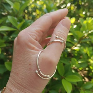 May include: A silver-colored ring with a unique design. The ring wraps around the thumb and features two small, spherical accents. The ring is worn on a hand with a blurred green background.