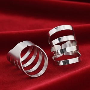May include: Two silver-colored finger rings. One ring has a cage-like design with four bands. The other ring has a stacked design with five bands. The rings are displayed on a red fabric surface.
