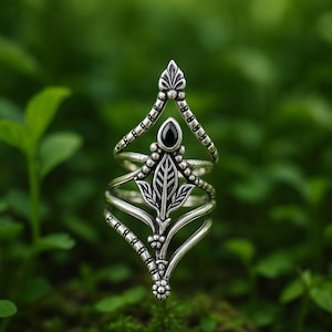 Op de afbeelding: Een zilverkleurige ring met een sierlijk, op de natuur geïnspireerd ontwerp. De ring heeft een centraal bladmotief, een zwarte edelsteen en delicate, met kralen bezette en gebogen accenten. Het ontwerp van de ring is symmetrisch en gedetailleerd, tegen een vage groene achtergrond.