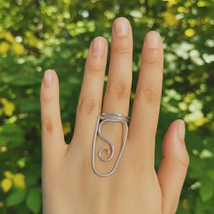 May include: A silver-colored ring with a unique design. The ring features a large, open oval shape with a swirled design inside. The ring is worn on a hand, with a blurred green background.