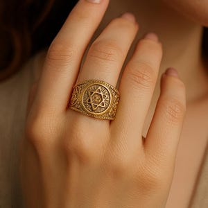 Op de afbeelding: Een goudkleurige ring met een Davidster-ontwerp. De ring heeft een ingewikkeld patroon rond de ster en op de band. De ring wordt aan een vinger gedragen.