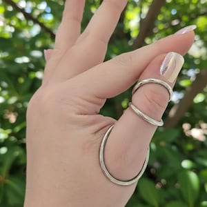May include: Three silver rings are worn on a thumb. One ring is at the base of the thumb, another is in the middle, and the third is at the tip. The rings are simple and minimalist in design. The background is blurred green.