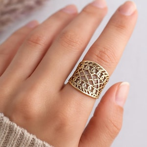 Op de afbeelding: Een goudkleurige ring met een open ontwerp met een geometrisch patroon van elkaar kruisende lijnen en kleine cirkelvormige vormen. De ring is breed en bedekt een aanzienlijk deel van de vinger.