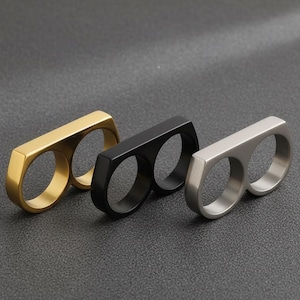 May include: Three double-finger rings in gold, black, and silver. Each ring has a rectangular bar connecting two circular bands. The rings are displayed on a dark gray surface, showcasing their minimalist design.