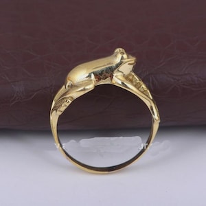 Könnte beinhalten: Ein goldfarbener Ring mit einem detaillierten Froschdesign. Der Frosch sitzt auf einer blattartigen Struktur, die das Band des Rings bildet. Der Ring hat eine polierte Oberfläche.