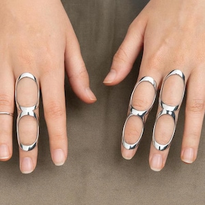 Op de afbeelding: Zilverkleurige vingerringen met een open ontwerp. De ringen hebben twee ovale openingen en worden op de vingers gedragen. Een uniek sieraad.