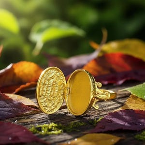 Könnte beinhalten: Ein goldener Medaillonring, geöffnet, um einen leeren Innenraum zu zeigen. Der Ring hat ein ovales Medaillon mit einem strukturierten Design auf der Innenseite. Der Ring ist vor einem Hintergrund aus bunten Herbstblättern platziert.