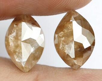 May include: Two marquise-cut, light brown diamonds. The gems are held between fingers, showcasing their faceted surfaces and warm, earthy tones. The diamonds have a pointed shape, with a symmetrical, elongated form.