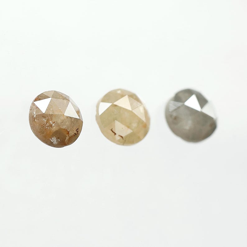 Op de afbeelding: Drie gefacetteerde, ovaalvormige diamanten in verschillende tinten bruin, geel en grijs. De edelstenen worden gepresenteerd tegen een witte achtergrond, waarbij hun unieke kleuren en slijpvormen worden getoond. De diamanten zijn waarschijnlijk voor het maken van sieraden.