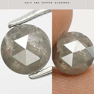 Op de afbeelding: Twee ronde zout- en peperdiamanten worden getoond. De diamanten zijn grijs met witte geometrische facetten. De diamanten worden vastgehouden door een pincet en een vinger. De tekst "SALT AND PEPPER DIAMONDS" staat bovenaan.
