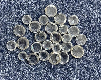 Könnte beinhalten: Eine Sammlung klarer, facettierter Edelsteine unterschiedlicher Größe. Die Edelsteine sind rund mit mehreren Facetten, die das Licht reflektieren. Sie sind auf einer strukturierten, dunkelblauen Oberfläche verstreut.