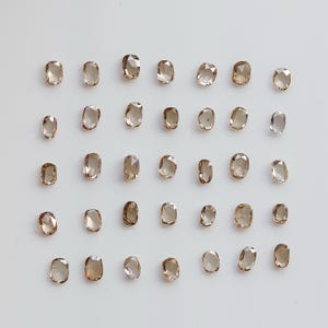 May include: A top-down view of numerous oval-shaped, light brown gemstones laid out in a grid. The gems are translucent, with differing shades of brown. Some appear clearer than others. The background is a plain white surface.