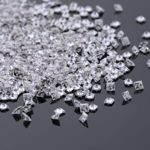 Könnte beinhalten: Eine Streuung kleiner, klarer, quadratisch geschliffener Edelsteine. Die Edelsteine sind hochreflektierend, fangen das Licht ein und erzeugen einen funkelnden Effekt. Die Steine liegen auf einer dunklen, reflektierenden Oberfläche.