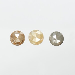 Op de afbeelding: Drie ronde, gefacetteerde diamanten in tinten bruin, geel en grijs. Elke diamant heeft een unieke kleur en interne structuur, die natuurlijke variaties laten zien. De diamanten zijn geplaatst tegen een effen witte achtergrond.