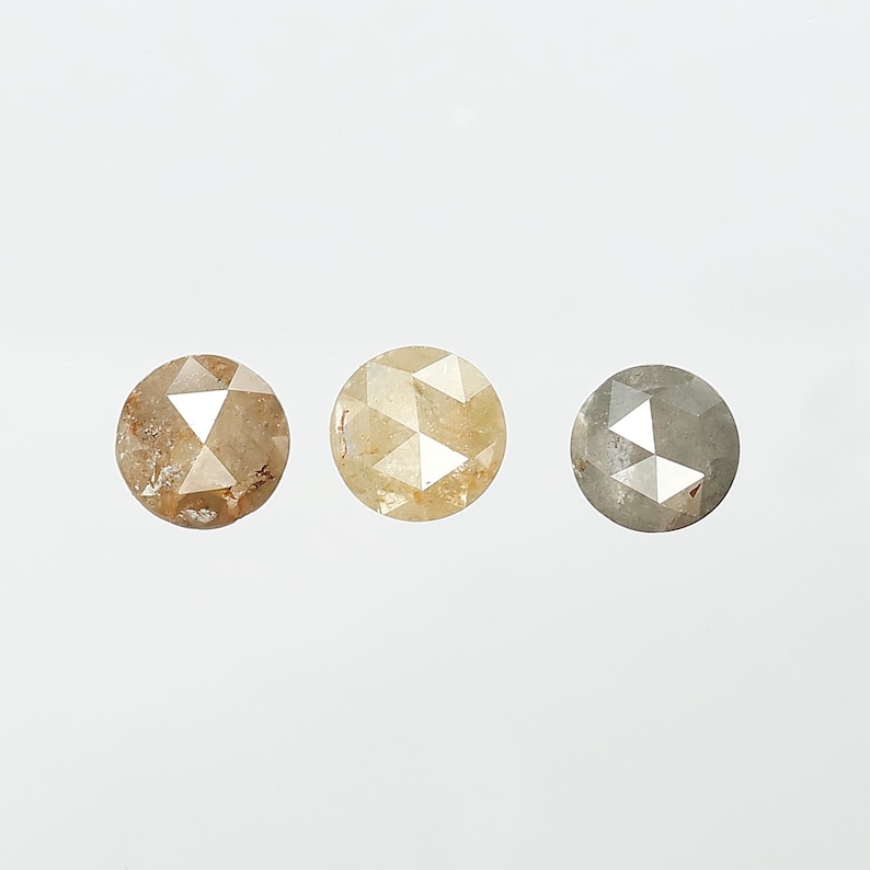 Op de afbeelding: Drie ronde, gefacetteerde diamanten in tinten bruin, geel en grijs. Elke diamant heeft een unieke kleur en interne structuur, die natuurlijke variaties laten zien. De diamanten zijn geplaatst tegen een effen witte achtergrond.
