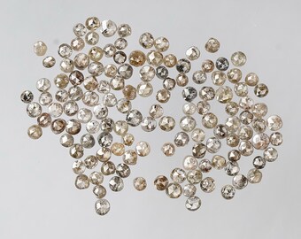 May include: A collection of small, round diamonds in various shades of brown, gray, and clear. The diamonds are scattered across a light-colored surface, each with multiple facets that catch the light.