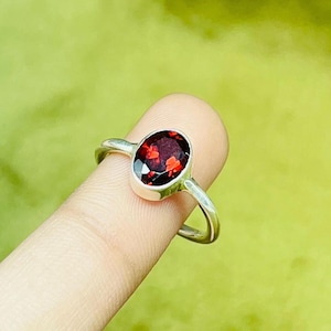 Op de afbeelding: Een zilveren ring met een dieprode ovale edelsteen. De edelsteen is gezet in een eenvoudige, elegante band. De ring wordt gepresenteerd tegen een zachte groene achtergrond, die de levendige kleur van de steen benadrukt.