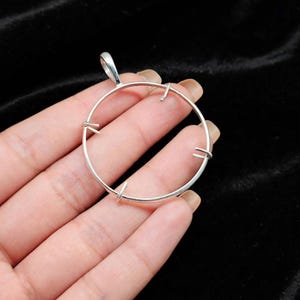 May include: A circular silver pendant setting featuring a thin metal frame with four small prongs extending inward from the perimeter. The pendant includes a small integrated bail at the top for attachment to a chain or cord, showcasing a metallic finish.
