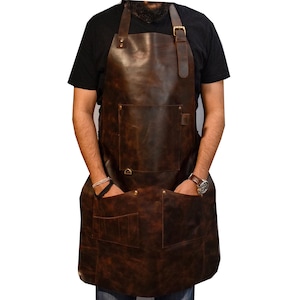 May include: A brown leather apron with multiple pockets and adjustable straps. The apron has a large front pocket, two lower pockets, and a small pocket on the chest. The straps are secured with brass buckles. The apron is designed for protection and utility.