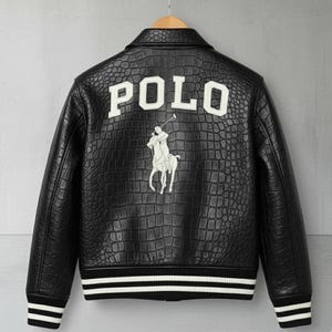 Puede incluir: Chaqueta de cuero negro con textura de cocodrilo, con la palabra "POLO" en letras blancas en la espalda. Un logotipo blanco de jugador de polo está debajo del texto. La chaqueta tiene un ribete a rayas blanco y negro en los puños y el dobladillo.