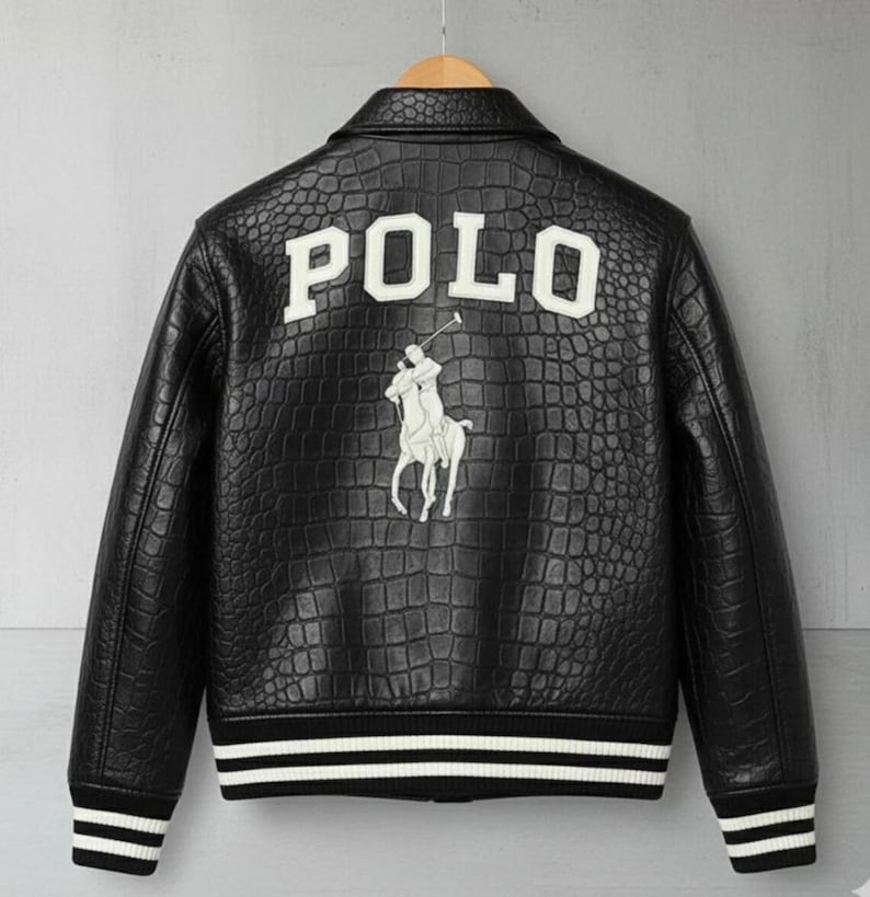 May include: Black leather jacket with a crocodile texture, featuring the word "POLO" in white block letters on the back. A white polo player logo is below the text. The jacket has a black and white striped trim on the cuffs and hem.