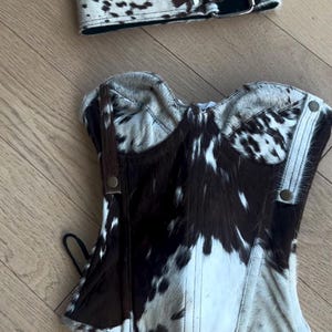 May include: A cowhide corset and matching belt. The corset is a structured top with a brown and white spotted pattern. The belt is wide with a silver buckle. The set is made of genuine leather.