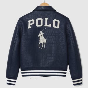 Puede incluir: Chaqueta de cuero azul marino con efecto de piel de cocodrilo. La parte trasera presenta la palabra "POLO" en letras blancas sobre un logotipo de jugador de polo blanco. Rayas blancas adornan los puños y el dobladillo.