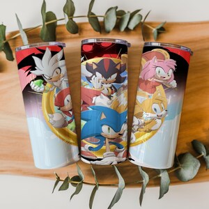 May include: Three cylindrical metal tumblers featuring colorful graphic prints of Sonic the Hedgehog characters including Silver, Knuckles, Shadow, Sonic, Amy Rose, and Tails. Each tumbler has a silver-toned rim and a smooth, glossy finish.