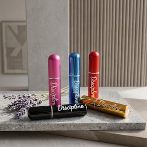 May include: A collection of colorful perfume atomizers. The atomizers are in various colors, including pink, blue, red, black, and gold. Each atomizer has the word "Discipline" printed on it in a cursive font. The atomizers are displayed on a gray surface.